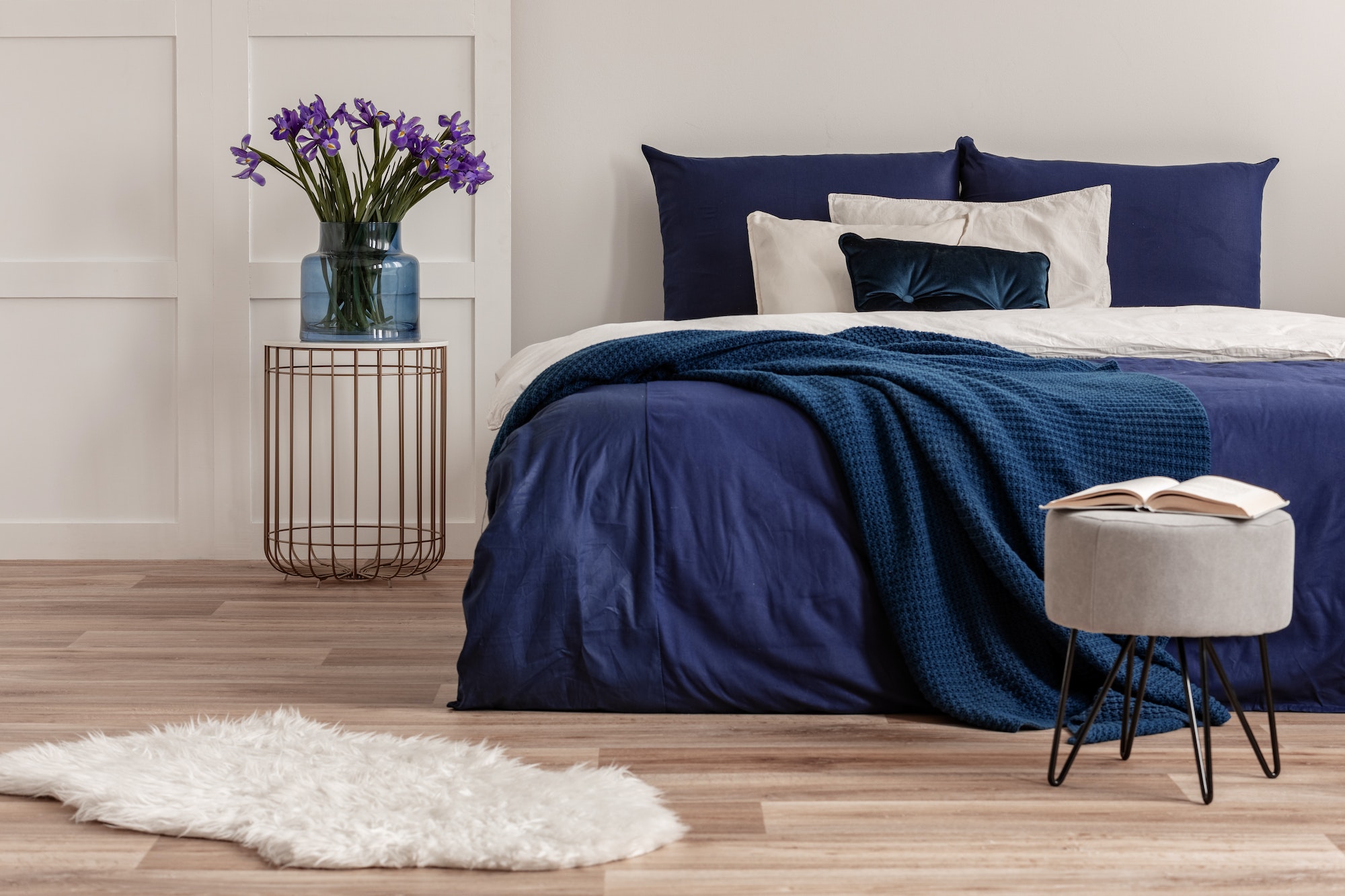 Purple flowers in blue glass vase on stylish bedside table next to king size bed